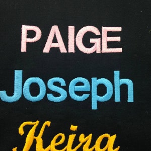 May include: Three names are embroidered on a black fabric background. The names are Paige in pink, Joseph in blue, and Keira in yellow.