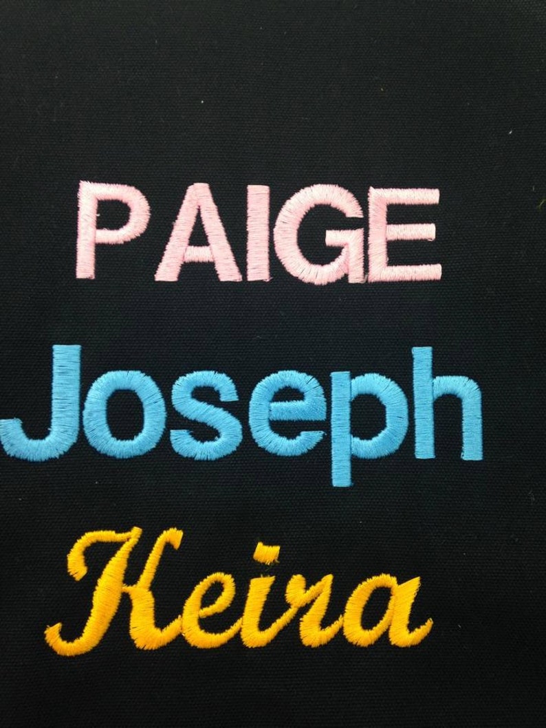 May include: Three names are embroidered on a black fabric background. The names are Paige in pink, Joseph in blue, and Keira in yellow.