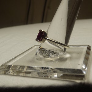 May include: A silver ring with a dark purple gemstone set in a prong setting. The ring is displayed on a clear ring holder.