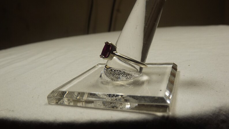 May include: A silver ring with a dark purple gemstone set in a prong setting. The ring is displayed on a clear ring holder.