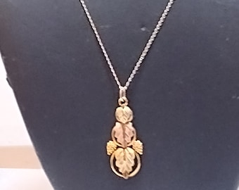 Pode incluir: Um colar de corrente de prata com um pingente de ouro que apresenta um grupo de folhas em tons de verde, amarelo e vermelho.