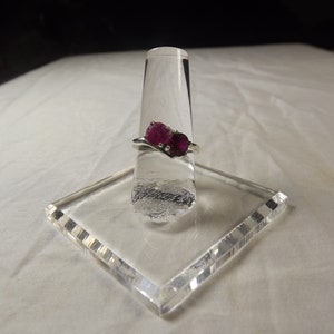 May include: A silver ring featuring two oval-cut, deep red gemstones. The ring is displayed on a clear, angled stand, showcasing its design. The gemstones are the focal point, set against the silver band.