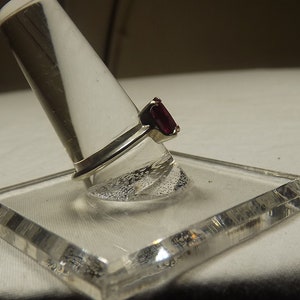 May include: A silver ring with a dark red rectangular gemstone. The ring is displayed on a clear plastic ring holder.