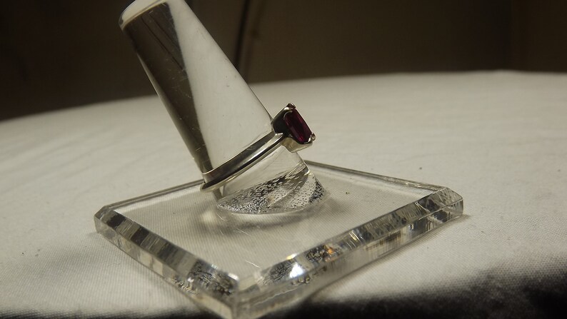 May include: A silver ring with a dark red rectangular gemstone. The ring is displayed on a clear plastic ring holder.