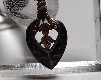 Pode incluir: Um pingente de coração preto com um design de folhas de ouro e prata. O pingente é feito de metal e tem um acabamento de esmalte preto.