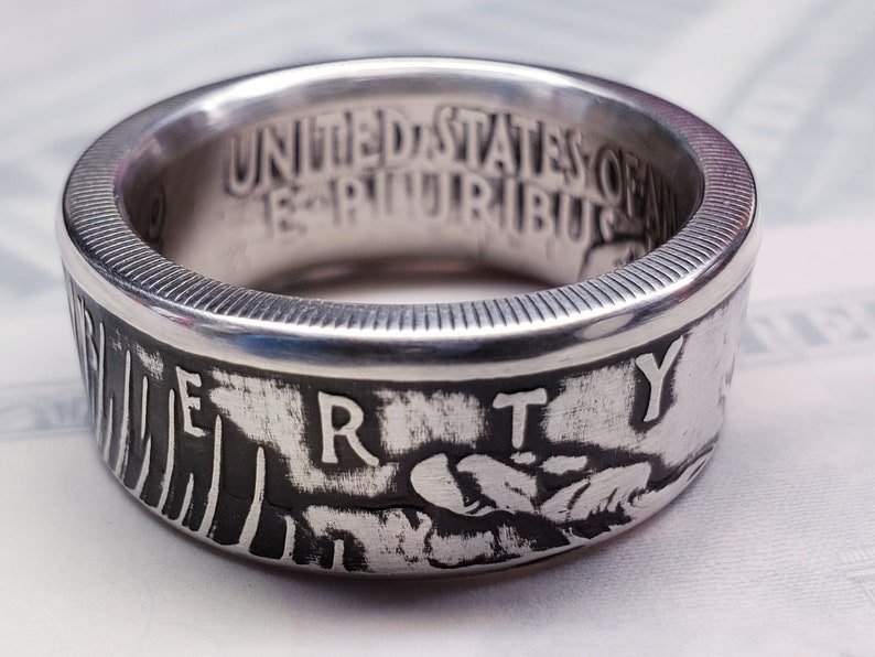 May include: Silver ring made from a United States coin. The ring features the words "United States of America" and "E Pluribus Unum" on the inside of the band. The outside of the band features the word "Liberty" and a design of an eagle's head.
