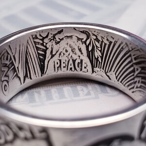 May include: A silver ring with the words "One Dollar" and "Peace" engraved on the inside. The ring also features an image of a woman's head and a branch of leaves.