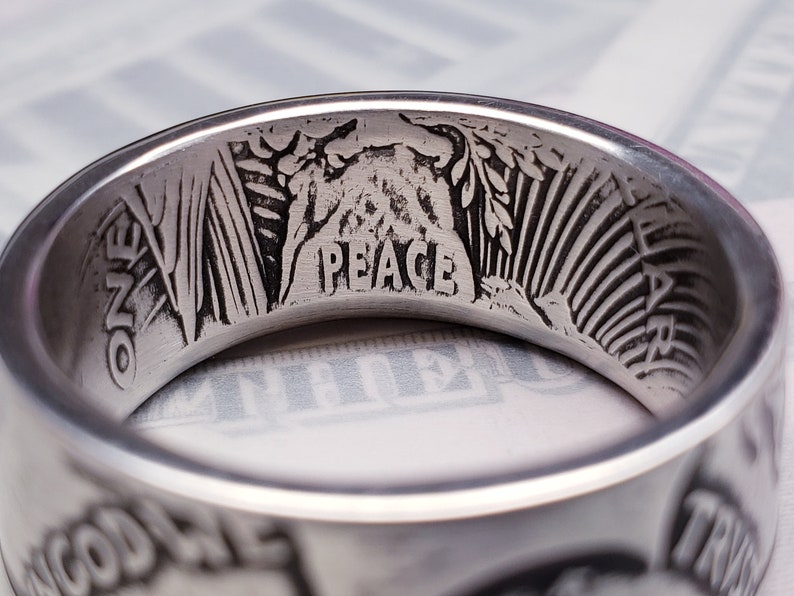 May include: A silver ring with the words "One Dollar" and "Peace" engraved on the inside. The ring also features an image of a woman's head and a branch of leaves.