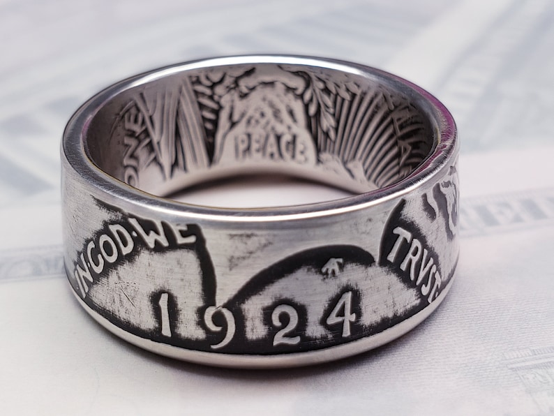May include: A silver ring made from a 1924 Peace Dollar coin. The ring features the inscription "In God We Trust" and the year "1924".