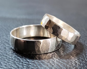 Puede incluir: Dos alianzas de boda de plata martilladas. Las alianzas son lisas y tienen una textura martillada. Las alianzas están apiladas una encima de la otra.