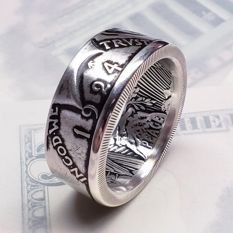 May include: A silver ring made from a 1924 Peace Dollar coin. The ring features the inscription "IN GOD WE TRUST" and "PEACE" on the side. The ring is made from a real coin and is a unique piece of jewelry.