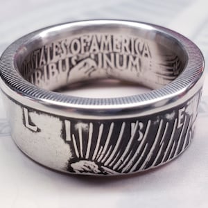 May include: Silver ring made from a US half dollar coin. The ring features the inscription "United States of America E Pluribus Unum" on the inside of the band and a depiction of Lady Liberty on the outside.