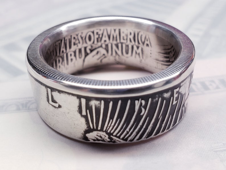 May include: Silver ring made from a US half dollar coin. The ring features the inscription "United States of America E Pluribus Unum" on the inside of the band and a depiction of Lady Liberty on the outside.