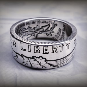May include: A silver ring made from a repurposed US half dollar coin. The ring features the words "LIBERTY" and a design of an eagle with outstretched wings.