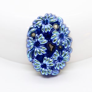 May include: A blue glass flower bead with a raised design. The bead has a metallic finish and is oval shaped.