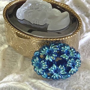 May include: A gold oval-shaped trinket box with an ornate design and a cameo of a woman's face. A blue flower-shaped brooch is sitting next to the box.