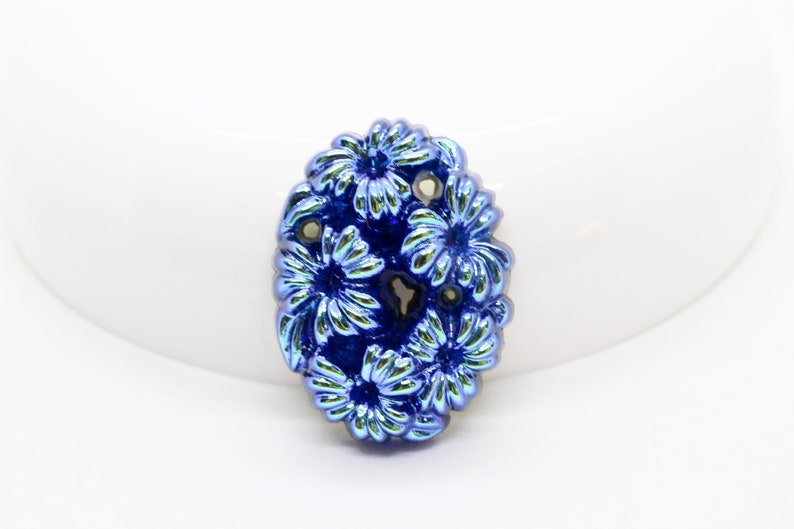 May include: A blue glass flower bead with a raised design. The bead has a metallic finish and is oval shaped.