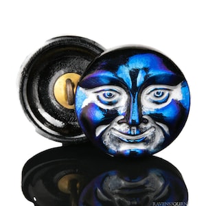 May include: A blue glass button with a white face and a smiling expression. The button is round and has a black backing.