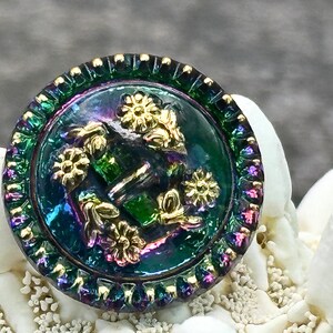 以下が含まれることがあります： 金色の縁取りと花柄のついた、虹色の緑色の丸いボタン。ボタンには、花と葉の浮き彫りのデザインが施されています。