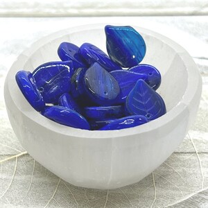 May include: A white bowl filled with blue glass leaf-shaped beads. The bowl is sitting on a white wooden surface.