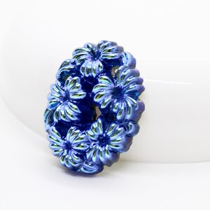 May include: A cluster of blue iridescent flowers in an oval shape. The flowers are made of metal and have a textured surface.
