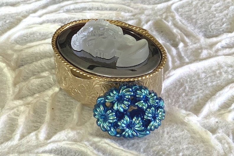 May include: A gold oval-shaped trinket box with an ornate design and a cameo of a woman's face. A blue flower-shaped brooch is sitting next to the box.