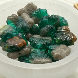 May include: A bowl filled with green and gray glass leaf-shaped beads. The beads are translucent and have a mottled appearance.