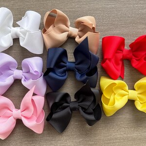 May include: Assortment of eight hair bows in various colors, including white, lavender, pink, yellow, red, navy blue, black, and beige. The bows are made of grosgrain ribbon and are tied with a center knot.