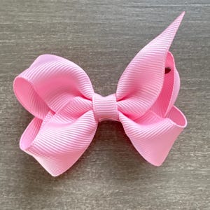 May include: A pink hair bow with a simple, classic design. The bow is made of a soft, textured fabric and is tied in a symmetrical knot.