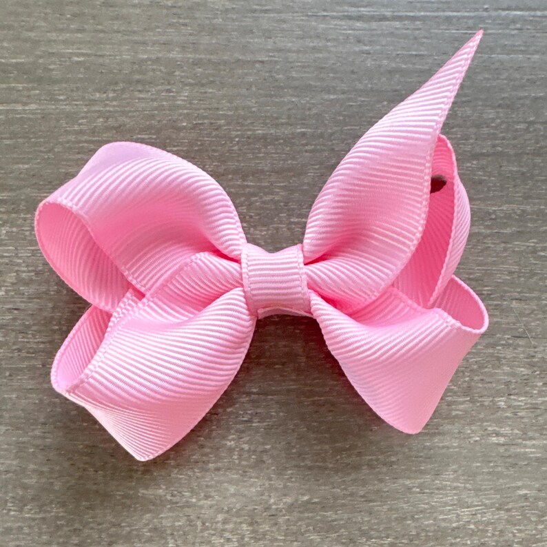 May include: A pink hair bow with a simple, classic design. The bow is made of a soft, textured fabric and is tied in a symmetrical knot.