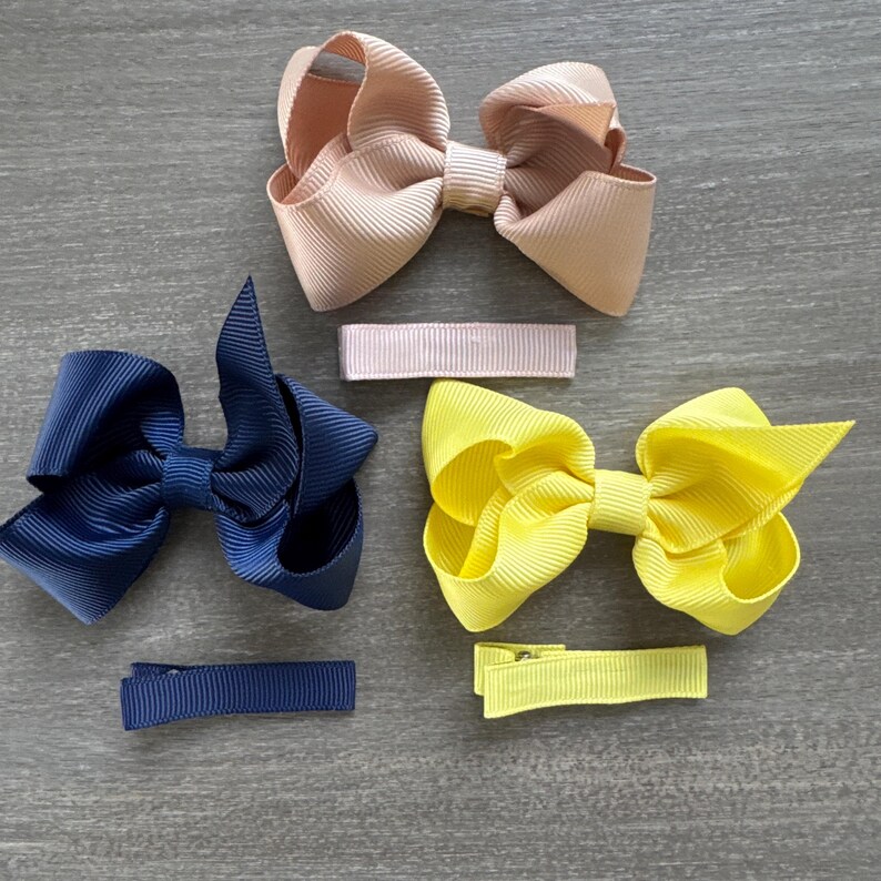 May include: Three hair bows with matching clips. The bows are made of grosgrain ribbon in navy blue, yellow, and blush pink. The clips are made of the same ribbon.