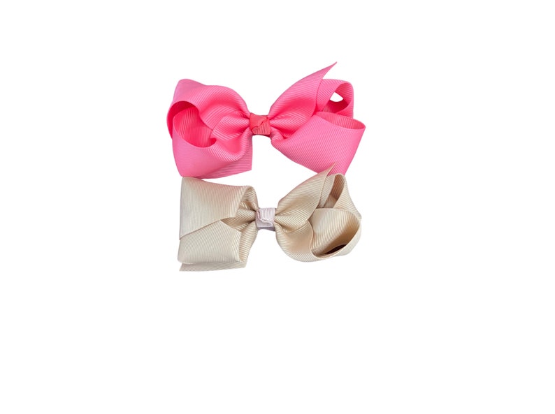 May include: Two hair bows, one pink and one beige, tied together with a white ribbon.