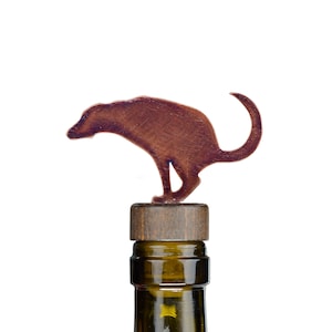 Puede incluir: Un tapón de botella de vino de metal de color cobre con forma de perro en posición de cuclillas. El tapón está encima de un corcho de botella de vino de madera.