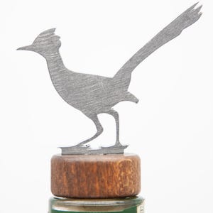 May include: A metal roadrunner bottle stopper. The silver roadrunner stands on a round, wooden base. The stopper is designed to fit on a bottle, adding a decorative touch. The background is white.