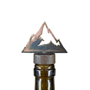 Könnte beinhalten: Ein Flaschenverschluss aus Metall in Form eines Gebirges. Der Verschluss ist aus Kupfer und hat eine dunkelbraune Holzbasis. Der Verschluss befindet sich auf einer grünen Glasweinflasche.