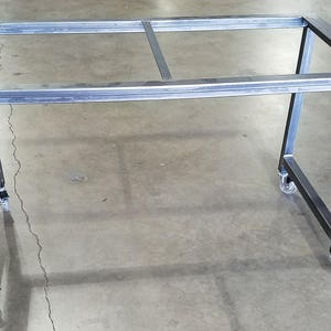 May include: A metal frame table with four wheels. The table is made of silver metal and has a rectangular shape. The table is on a gray concrete floor.