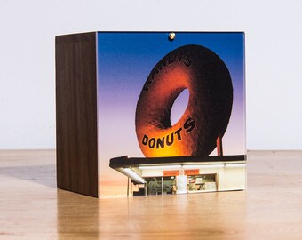 May include: A wooden box with a photo of a giant donut sign that says "Randy's Donuts". The sign is lit up and the sky is a gradient of purple, pink, and orange.