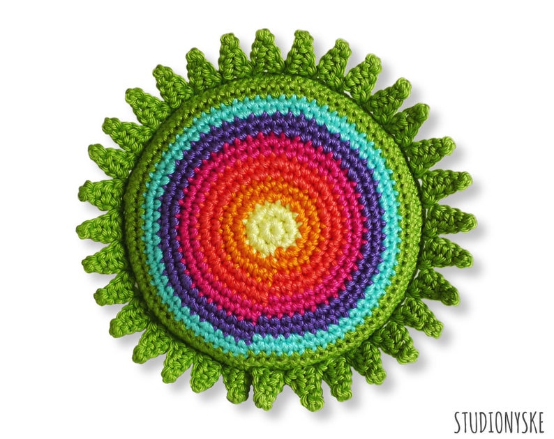 Puede incluir: Un sol de crochet colorido con un anillo exterior verde y un centro de arco&iacute;ris. El centro est&aacute; hecho de m&uacute;ltiples c&iacute;rculos conc&eacute;ntricos de diferentes colores, incluyendo rojo, naranja, amarillo, verde, azul, morado y rosa.