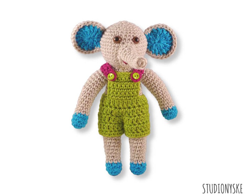 May include: A crocheted elephant toy wearing green dungarees with pink and yellow buttons. The elephant has blue ears and a brown nose.