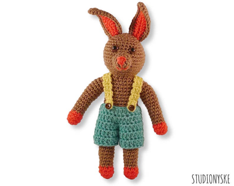 May include: A brown crocheted bunny wearing blue dungarees with yellow straps and orange shoes.