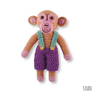 May include: A crocheted monkey toy wearing purple dungarees with green straps and yellow buttons. The monkey has a pink face and brown fur.