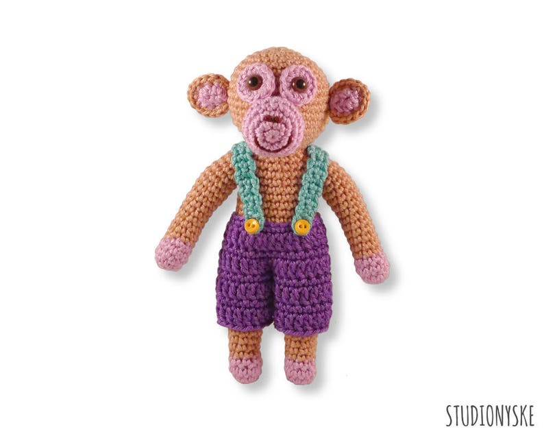 May include: A crocheted monkey toy wearing purple dungarees with green straps and yellow buttons. The monkey has a pink face and brown fur.