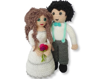 May include: A crocheted bride and groom doll set. The bride is wearing a white dress with a silver trim and holding a bouquet of pink and red flowers. The groom is wearing a white shirt, gray pants, and a turquoise bow tie.