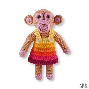 May include: A crocheted monkey doll wearing a colourful striped dress with a yellow bib and a pink button. The monkey has pink ears, a pink nose, and brown eyes.