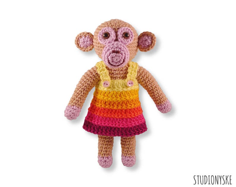May include: A crocheted monkey doll wearing a colourful striped dress with a yellow bib and a pink button. The monkey has pink ears, a pink nose, and brown eyes.