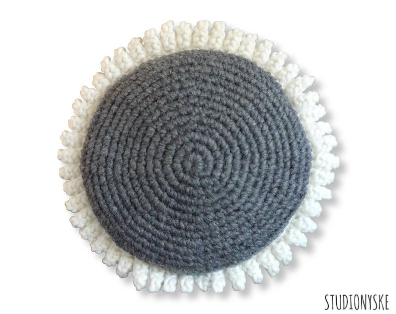 Puede incluir: Un coj&iacute;n redondo de crochet gris con un borde de crochet blanco. El coj&iacute;n est&aacute; hecho con un hilo texturizado y tiene un patr&oacute;n circular.