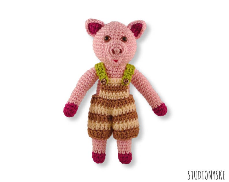 May include: A pink crocheted pig wearing brown and cream striped dungarees with green straps. The pig has a pink snout and pink ears.