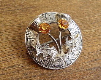Könnte beinhalten: Silberfarbene Brosche mit Distel-Design und zwei orange-gelben Edelsteinen. Die runde Brosche weist detaillierte Gravuren von Blättern und Distelköpfen auf. Die Edelsteine sind rund und in der Mitte der Distelblüten platziert.