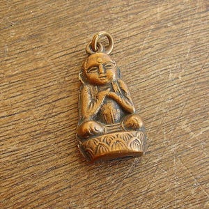 May include: A small, bronze-colored pendant depicting a seated Buddha figure. The Buddha is in a meditative pose, with hands clasped in front. The pendant has a loop at the top for attaching to a chain or cord. The base of the pendant has a decorative floral design.
