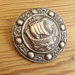 May include: A circular silver-toned metal brooch featuring a relief design of a Viking longship with a single sail on stylized waves. The central ship motif is surrounded by a border of raised circular dots and intricate knotwork patterns.